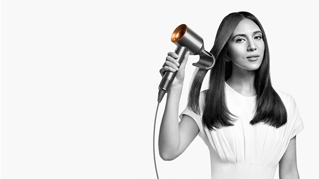 Een vrouw gebruikt een haardroger. De haardroger is zilverkleurig met een oranje accent. De achtergrond is wit.