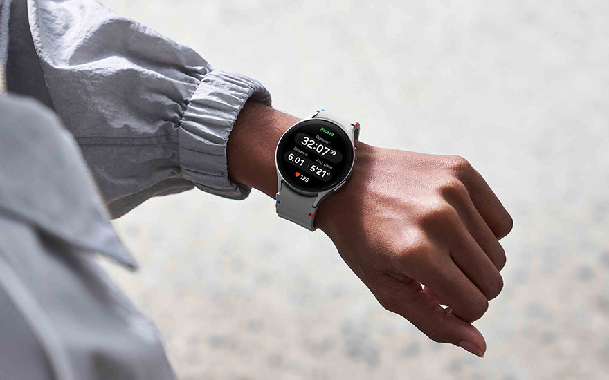 Persoon draagt zwarte Samsung-smartwatch met ronde wijzerplaat die stappen en hartslag toont, aan pols met grijze sportkleding in buitenomgeving.