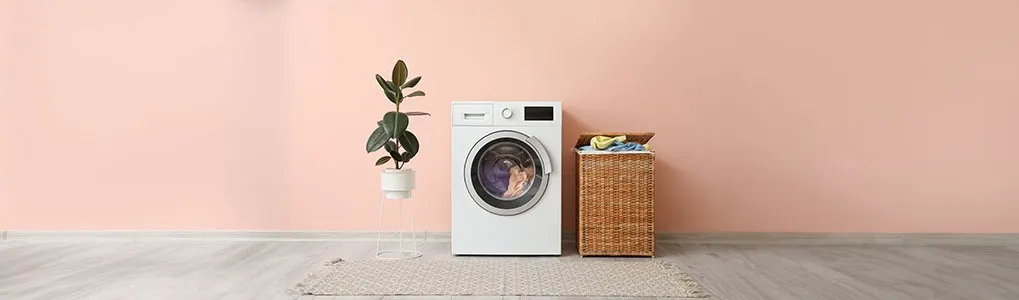 A white washing machine sits next to a wicker laundry basket on a gray rug. A potted plant is nearby, against a peach wall.