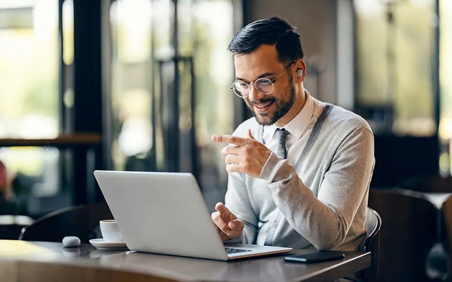 Man with glasses and earbuds smiles during video call on laptop in café, pointing at screen, coffee and phone on table.