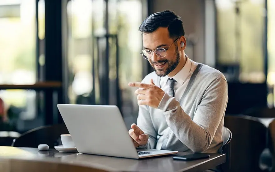 Man with glasses and earbuds smiles during video call on laptop in café, pointing at screen, coffee and phone on table.