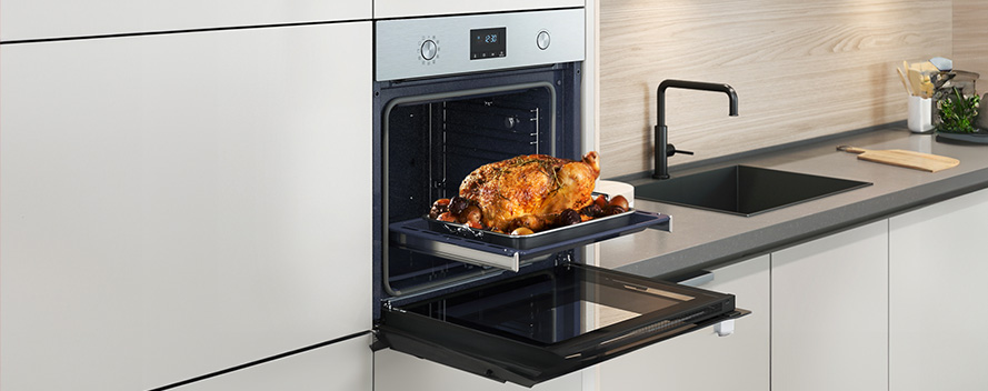 Roasted chicken on a tray in an oven with an open door in a kitchen with a sink, faucet and countertop.