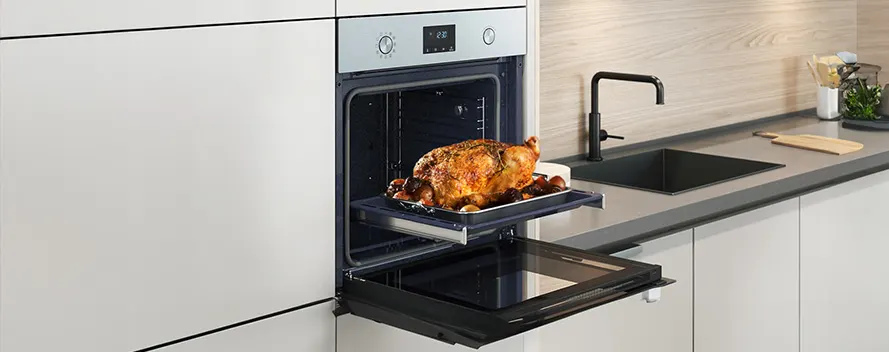 Roasted chicken on a tray in an oven with an open door in a kitchen with a sink, faucet and countertop.