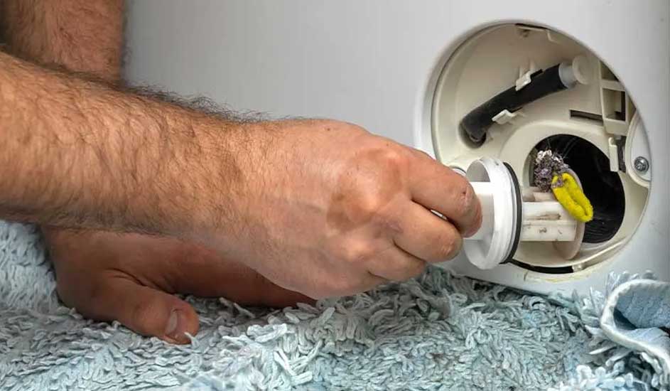 Een hand draait het filter van een wasmachine open voor onderhoud of reiniging. De wasmachine staat op een lichtblauw kleed.