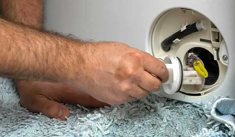 Een hand draait het filter van een wasmachine open voor onderhoud of reiniging. De wasmachine staat op een lichtblauw kleed.