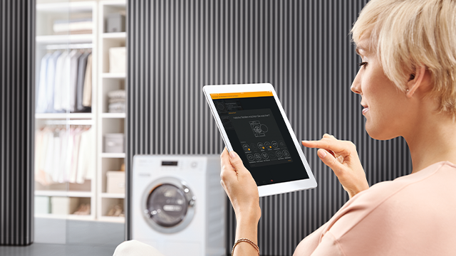 Vrouw gebruikt tablet in een kamer met een wasmachine en kledingkast tegen een gestreepte muur.