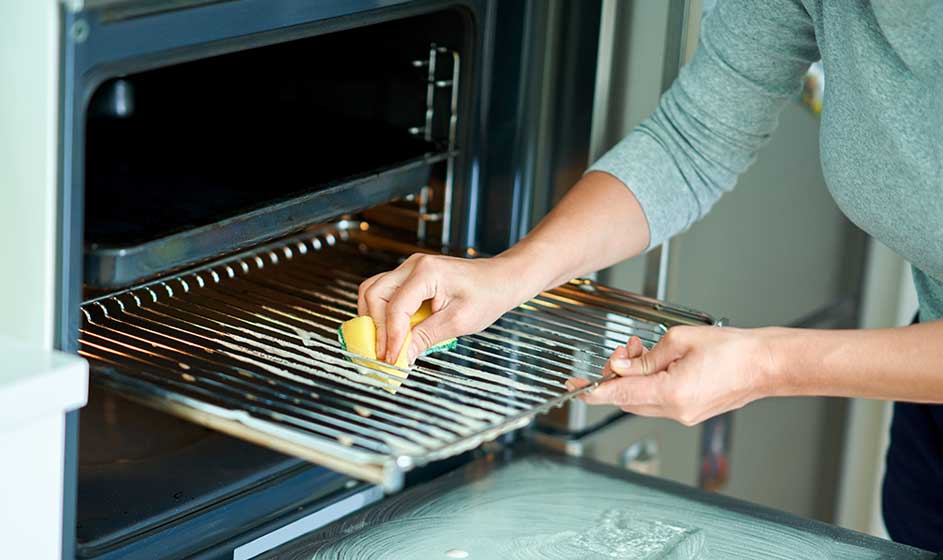 Someone cleans the oven rack with a sponge while the door stands open; the scene feels homely, tidy and practical.