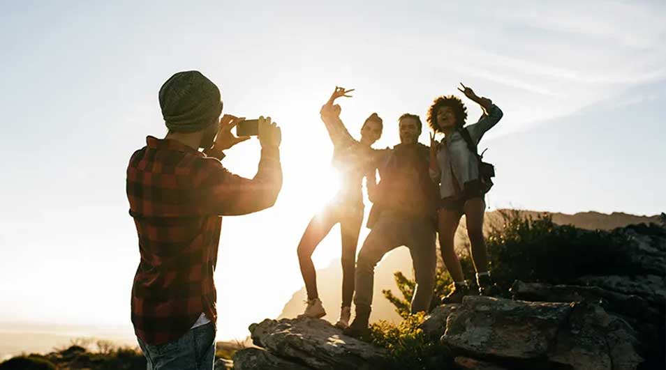 Vier vrienden poseren vrolijk op een rots bij zonsondergang terwijl een ander met een smartphone een foto maakt. Merk van de telefoon is niet zichtbaar.