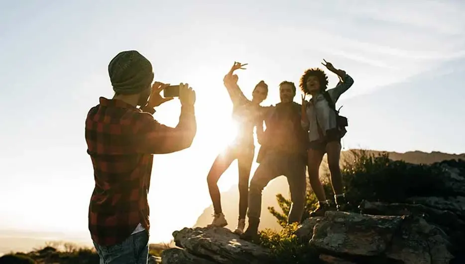 Vier vrienden poseren vrolijk op een rots bij zonsondergang terwijl een ander met een smartphone een foto maakt. Merk van de telefoon is niet zichtbaar.