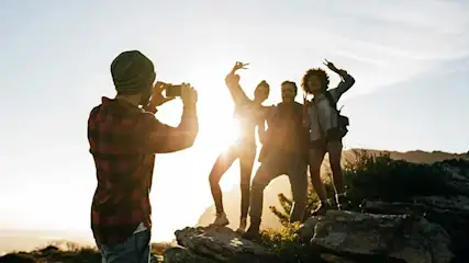 Friends pose on sunlit rocks as one person in a beanie photographs them, bright evening light outlining their playful silhouettes.