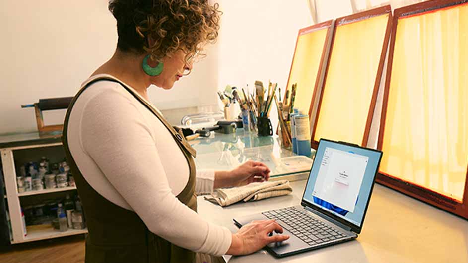 Een vrouw gebruikt een laptop op een bureau met penselen en schermen. Een kast staat aan de linkerkant.