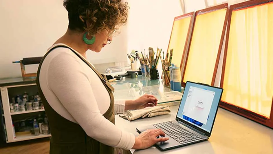 Een vrouw gebruikt een laptop op een bureau met penselen en schermen. Een kast staat aan de linkerkant.