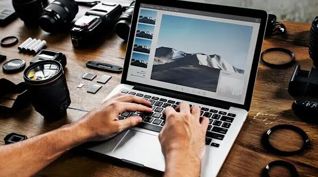 Handen typen op laptop met berglandschap op scherm, camera-uitrusting en lenzen rondom, fotografie werkplek setup