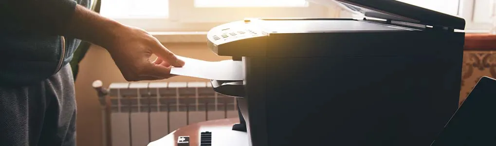 Een persoon gebruikt een laserprinter in een ruimte met ochtendlicht, terwijl hij een vel papier plaatst voor printen of scannen.