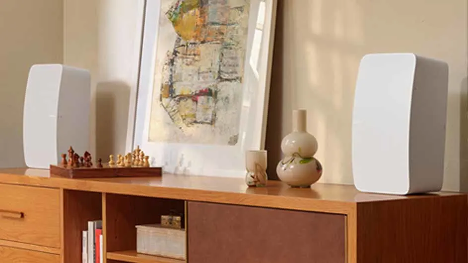 Twee witte speakers op een houten dressoir met een schilderij, een schaakspel en vazen.