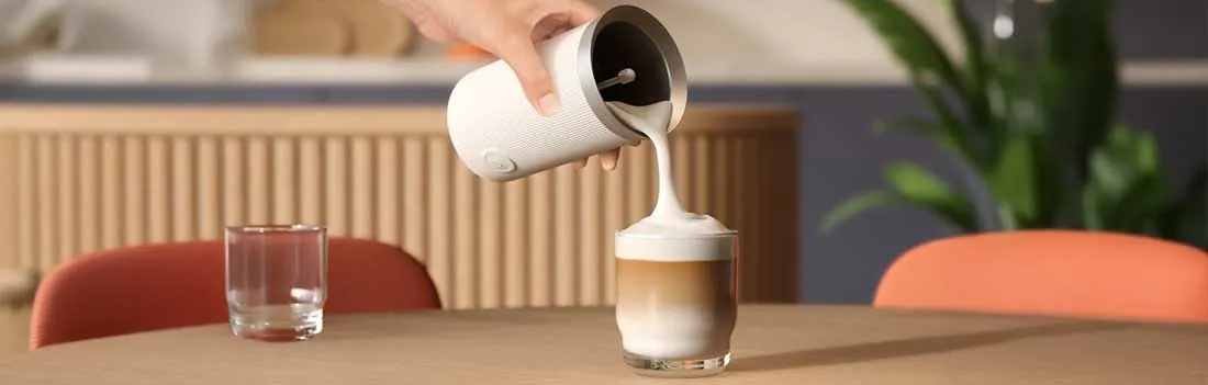 Een hand schenkt schuimende melk uit een witte opschuimer op een glas latte, op een tafel met glas water, stoel en plant erachter.