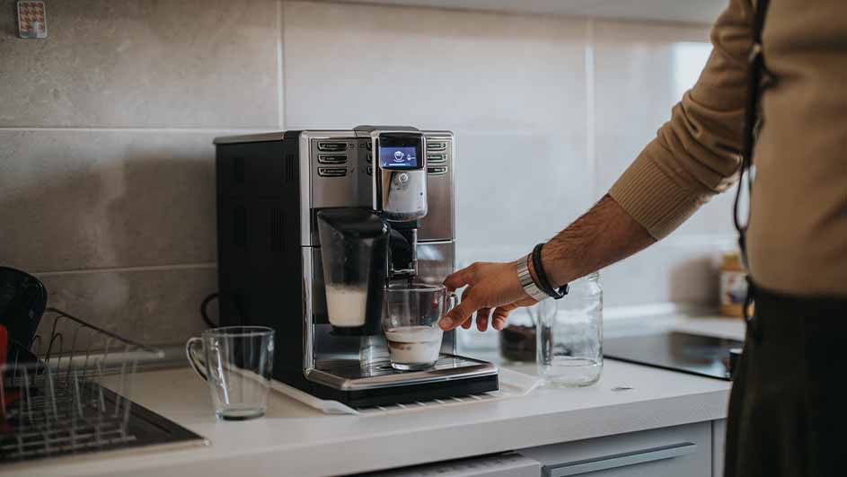 Person using a fully automatic espresso machine with milk frother to prepare a coffee drink in a kitchen.