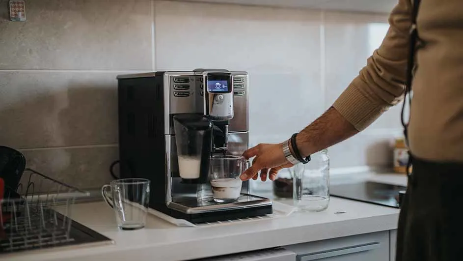 Person using a fully automatic espresso machine with milk frother to prepare a coffee drink in a kitchen.