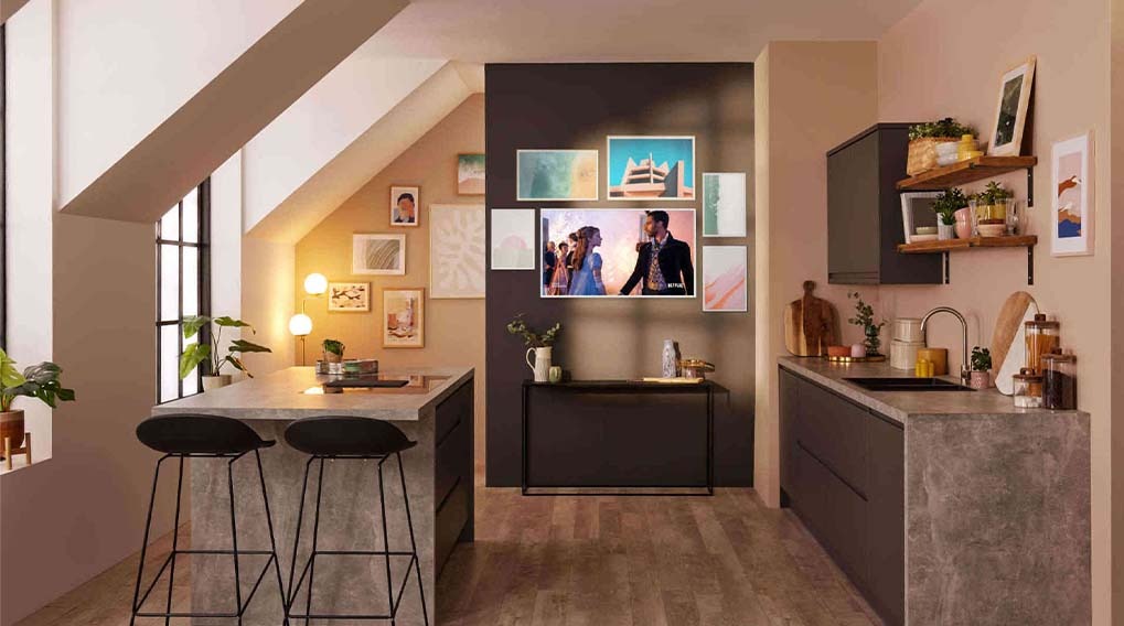 A modern kitchen setting with a TV mounted on the wall among several artworks, complemented by an island with two stools.