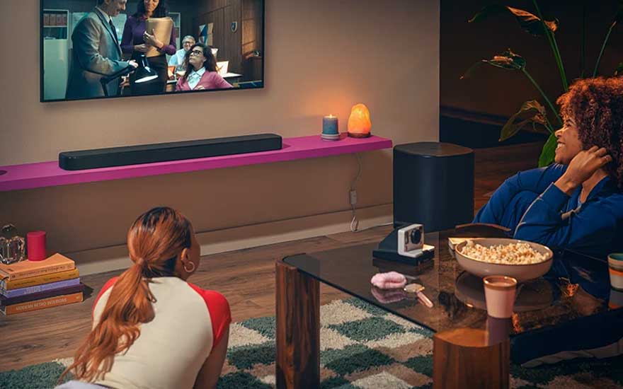 Twee vrouwen kijken film in sfeervol interieur met wand-tv, soundbar op roze plank, snacks, kaarsen en warme verlichting.