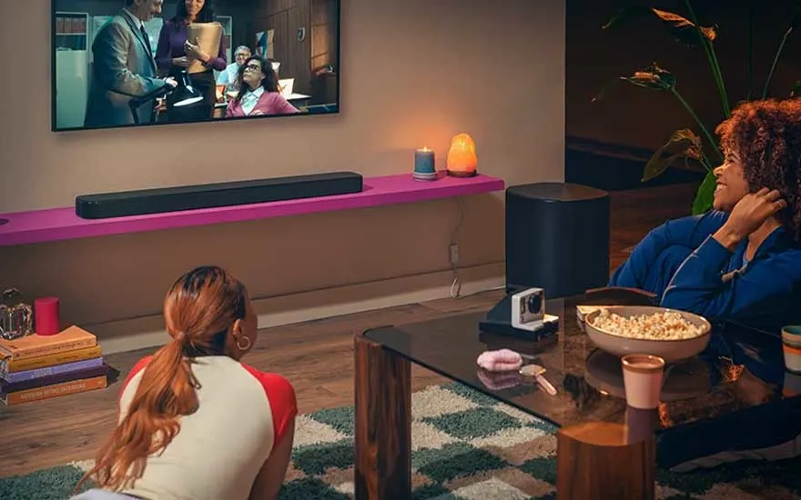Twee vrouwen kijken film in sfeervol interieur met wand-tv, soundbar op roze plank, snacks, kaarsen en warme verlichting.