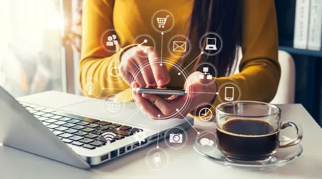 Persoon in gele trui gebruikt telefoon, met laptop en koffie in de buurt, omringd door digitale iconen.