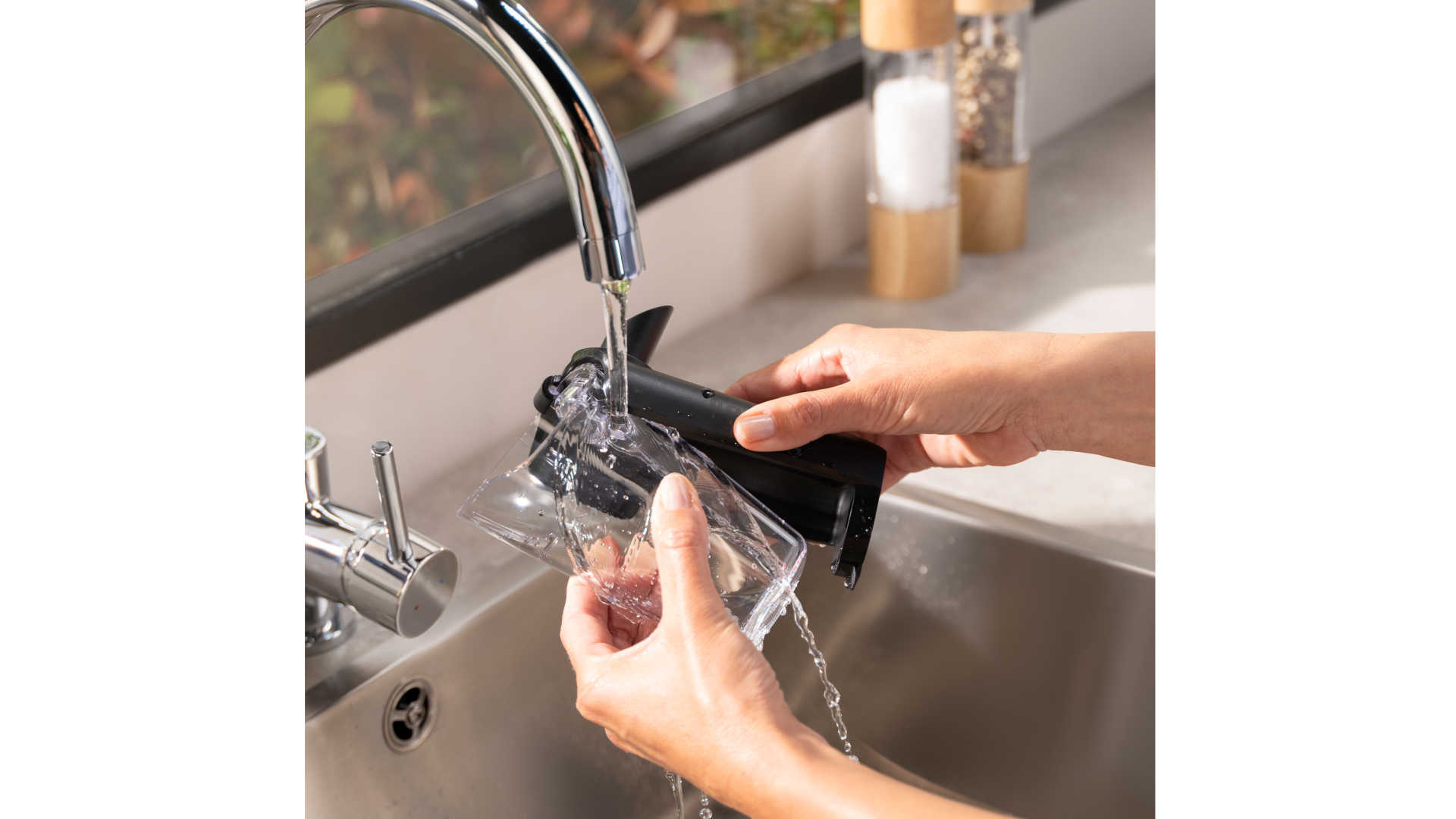 Handen spoelen een doorzichtige beker en een zwart onderdeel onder een zilveren kraan boven een spoelbak.