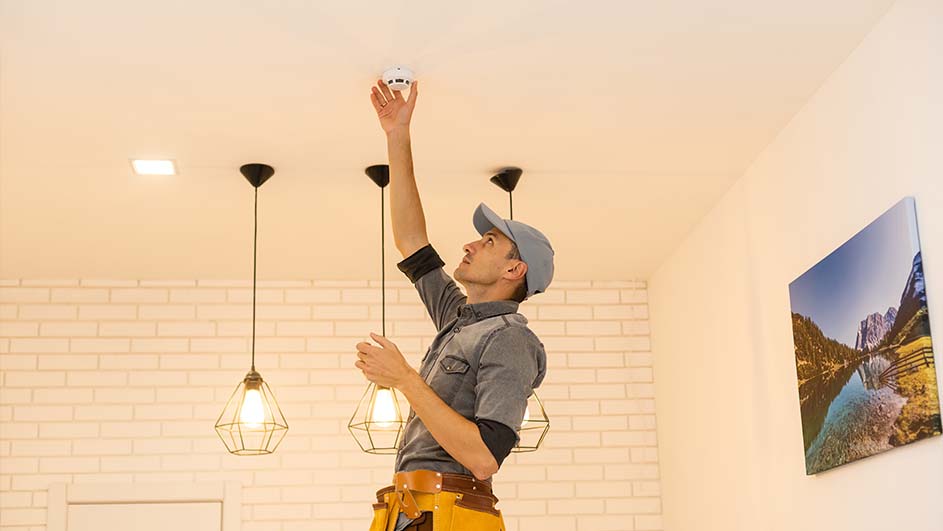 Een man met een gereedschapsgordel installeert een rookmelder aan het plafond, onder drie hanglampen, in een kamer met een schilderij.