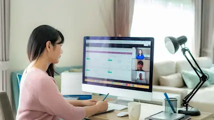 Een vrouw zit aan een bureau met een computer waarop een videovergadering te zien is, naast een lamp en kantoorartikelen.