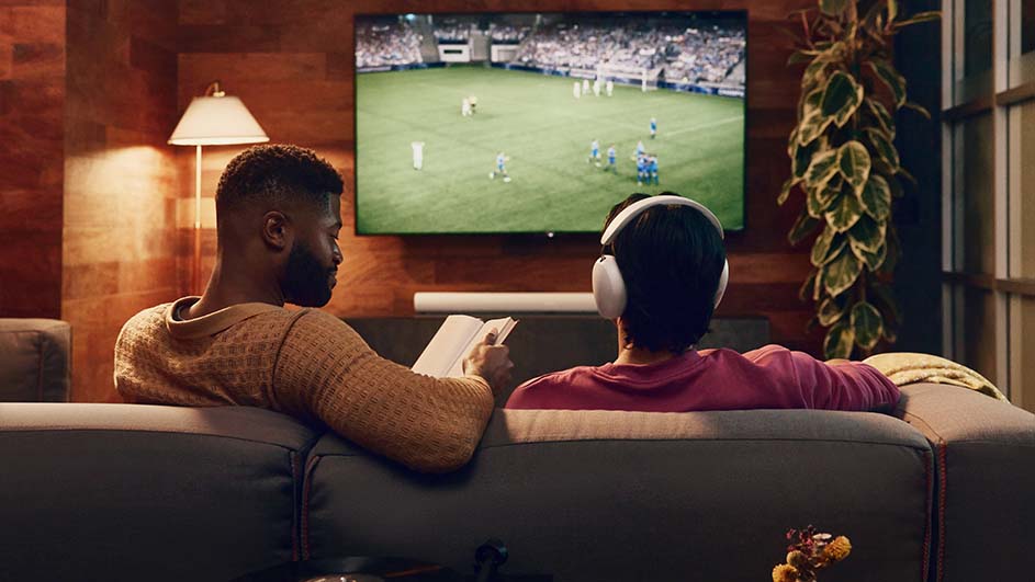 Twee mensen op een bank kijken naar voetbal op tv. Eén leest een boek, de ander draagt een koptelefoon. Een plant staat naast de tv.