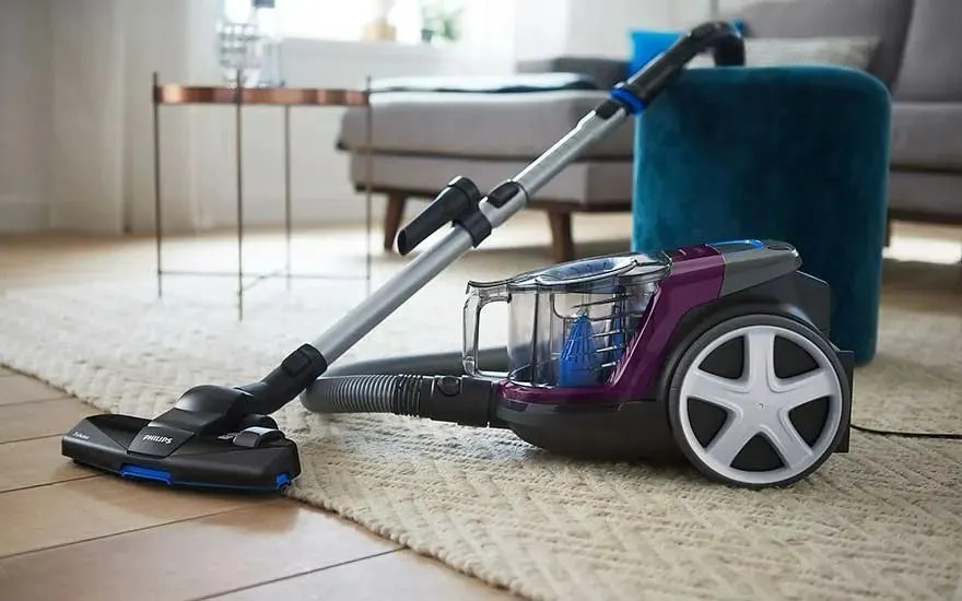 A black and purple Philips cylinder vacuum cleaner with transparent dust container is on a light rug in a modern living room with a sofa.
