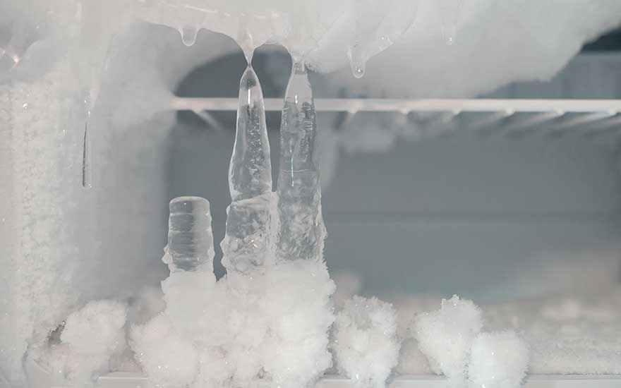 Interior of a freezer covered in heavy frost, with a thick layer of ice and long icicles hanging down from the top shelf and back wall.