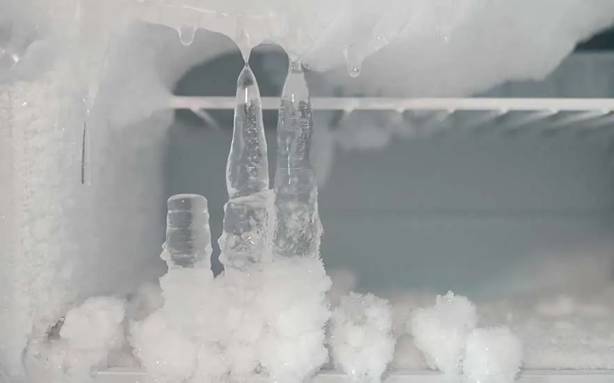 Interior of a freezer covered in heavy frost, with a thick layer of ice and long icicles hanging down from the top shelf and back wall.