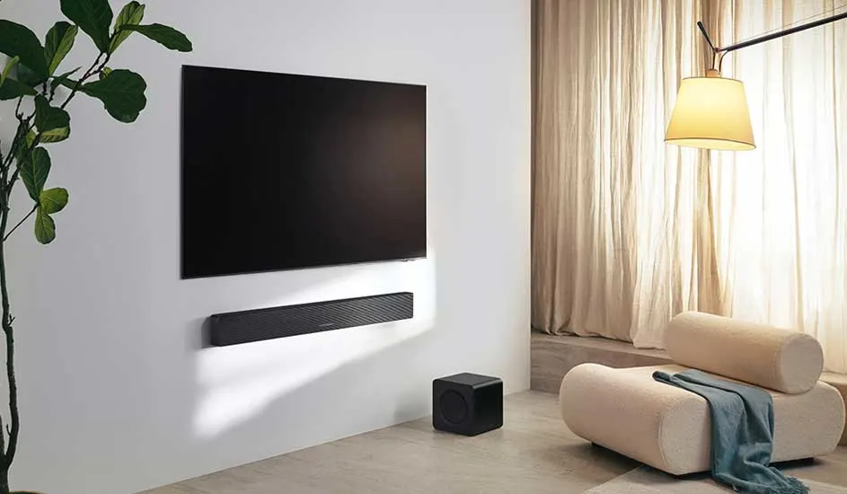 Een moderne woonkamer met een grote zwarte tv aan de muur, een soundbar eronder, een subwoofer op de grond en een stijlvolle beige stoel.