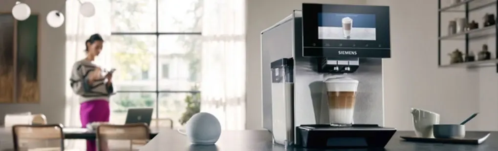 Een Siemens koffiemachine maakt een latte macchiato op een moderne keukentafel, met een kopje en smart speaker erbij.
