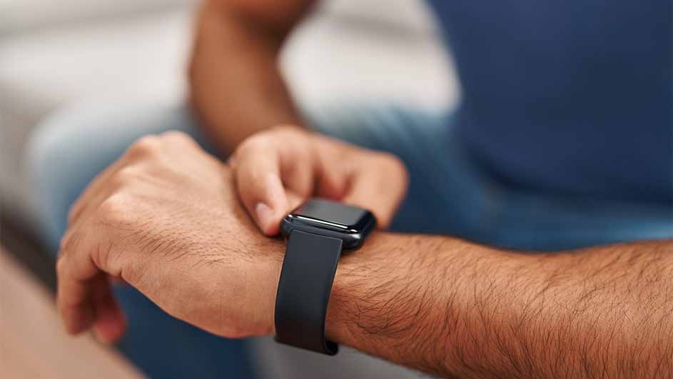 A person in a blue shirt looks at a black smart watch on their left wrist. The background is blurred.