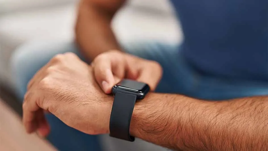 A person in a blue shirt looks at a black smart watch on their left wrist. The background is blurred.