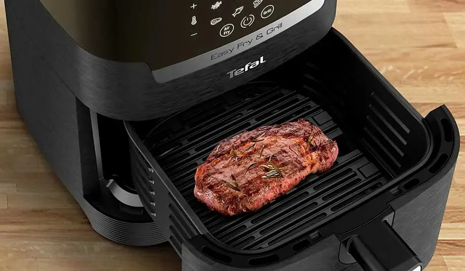 In a black Tefal Easy Fry & Grill air fryer lies a grilled piece of steak with herbs on the grill rack, on a wooden worktop.