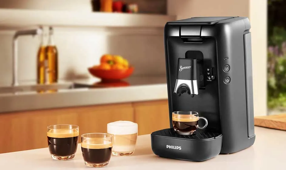 A black Philips coffee machine stands on the worktop, with three cups of coffee in front and a warm kitchen with fruit in the background.