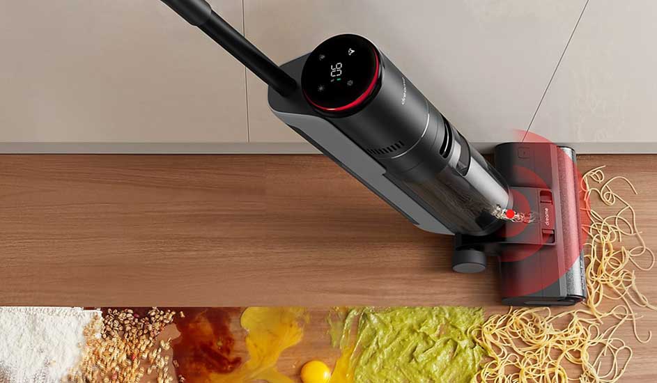 A cordless stick vacuum with a round display tackles a huge kitchen mess of flour, egg, sauce, noodles and veg on the wooden floor.