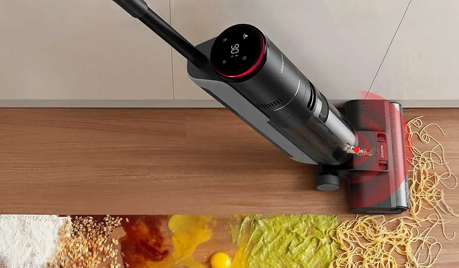 A cordless stick vacuum with a round display tackles a huge kitchen mess of flour, egg, sauce, noodles and veg on the wooden floor.