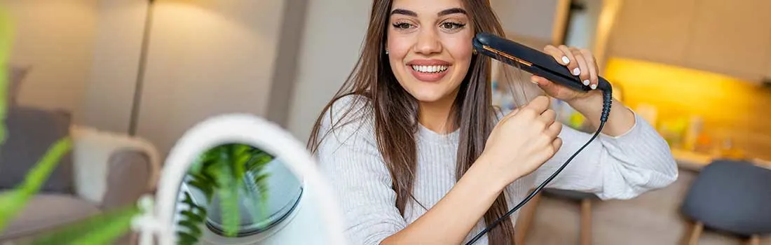 A smiling woman straightens her long hair with a flat iron at home, looking into a mirror in warm, soft lighting.