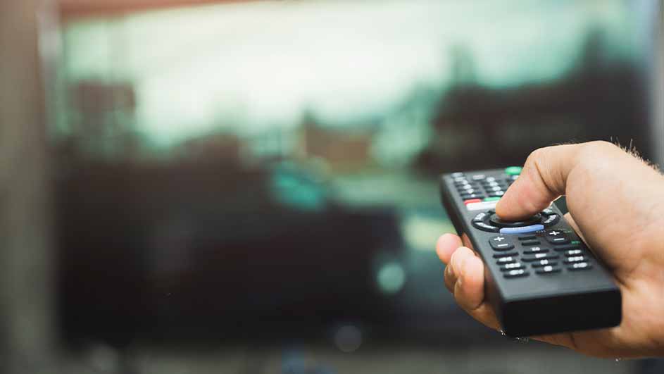 Een hand houdt een zwarte afstandsbediening vast voor een wazige tv, met groene, rode en witte knoppen.