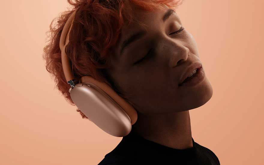 A person with short orange hair wears peach-coloured over-ear headphones, eyes closed in enjoyment against a soft apricot-toned background.