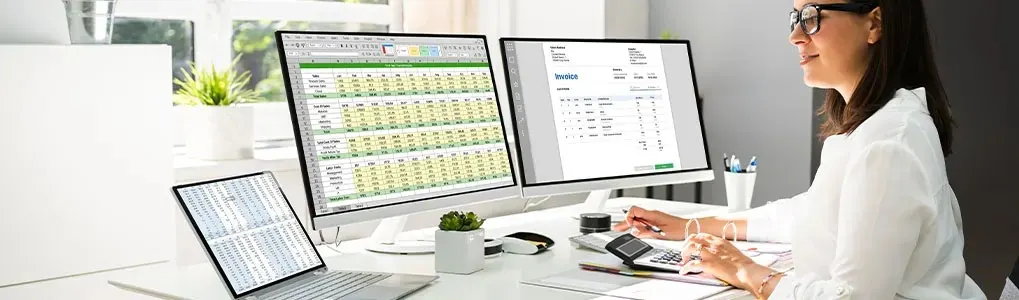 A person works at a desk with two monitors, a laptop, a calculator, and office supplies. The monitors display spreadsheets and invoices.