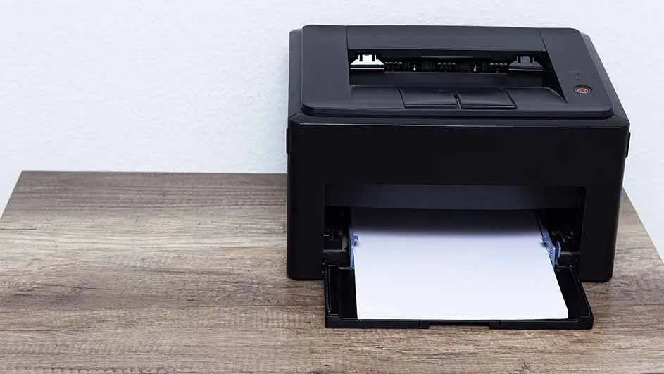 Een zwarte printer staat op een houten tafel tegen een witte achtergrond. Een lade met wit papier steekt uit de printer.