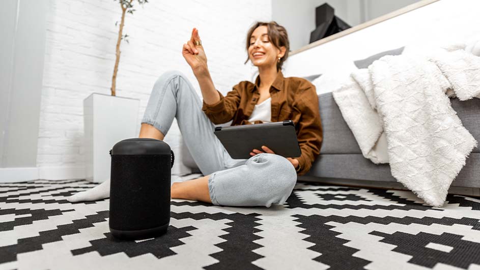 Vrouw zit op een zwart-wit kleed met een tablet, naast een zwarte speaker en grijze bank met witte deken.