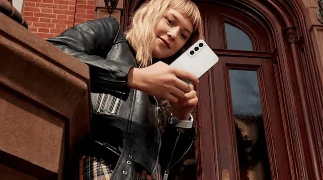 Een persoon houdt een witte Samsung smartphone vast en draagt een zwarte leren jas, staand voor een grote houten deur en een bakstenen muur.