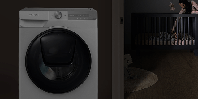 Een grijze Samsung wasmachine staat naast een openstaande deur naar een donkere kinderkamer met een wieg.