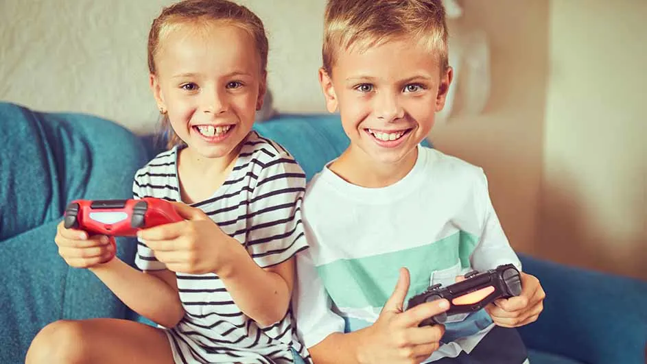 Twee kinderen zitten op een blauwe bank en lachen, terwijl ze gamecontrollers vasthouden.